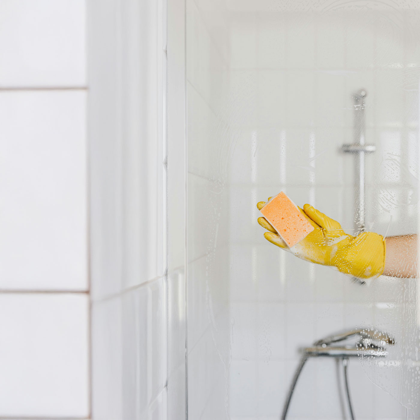 Ruka v žluté gumové rukavici čistí skleněnou stěnu sprchy houbičkou.